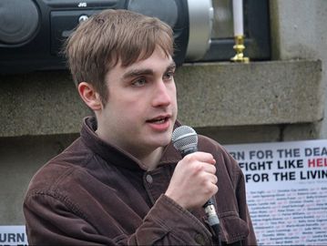 A white person holding and speaking into a microphone. Behind is a stone monument with posters attached.