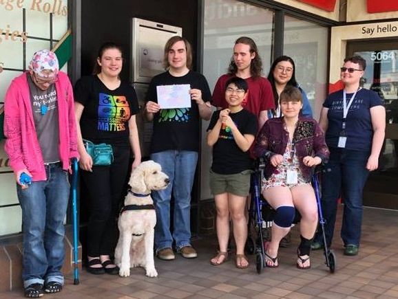 People standing outside and smiling for a group photo, some of them wearing disability and neurodiversity pride shirts and some have mobility aids, sunglasses, or a service dog with them. The person in the middle is holding a sign that says 